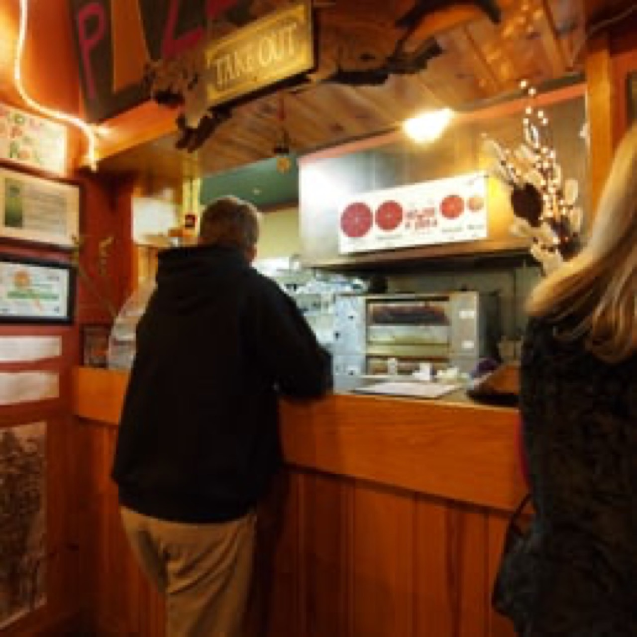 Customers ordering at a warm wood-paneled take-out counter with oven visible.